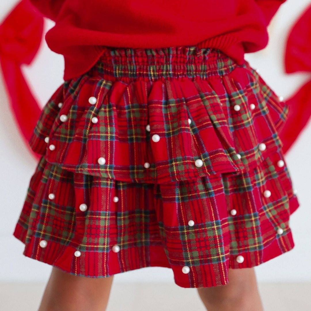 A close-up of a child wearing a red tiered plaid skirt with white pearl embellishments and a red knit top, photographed against a light background.