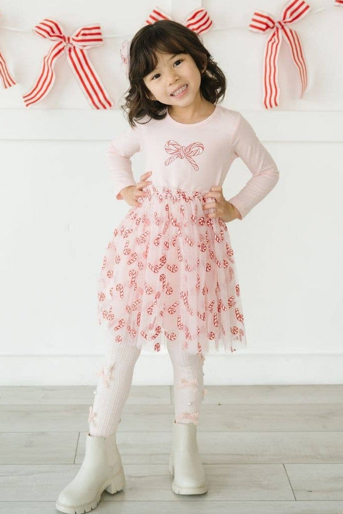 A young girl stands in a bright room wearing a light pink long-sleeve dress with a candy cane bow detail, a pink tulle skirt with candy cane embellishments, white leggings, and white boots. Red and white striped bunting is visible in the background.
