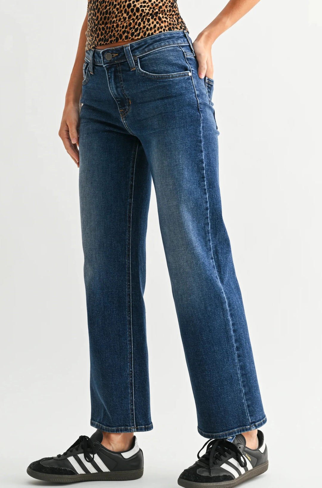 A model wearing a pair of medium-wash blue straight-leg jeans, styled with a leopard-print top and black sneakers, photographed against a light gray studio background, turned slightly to the side.