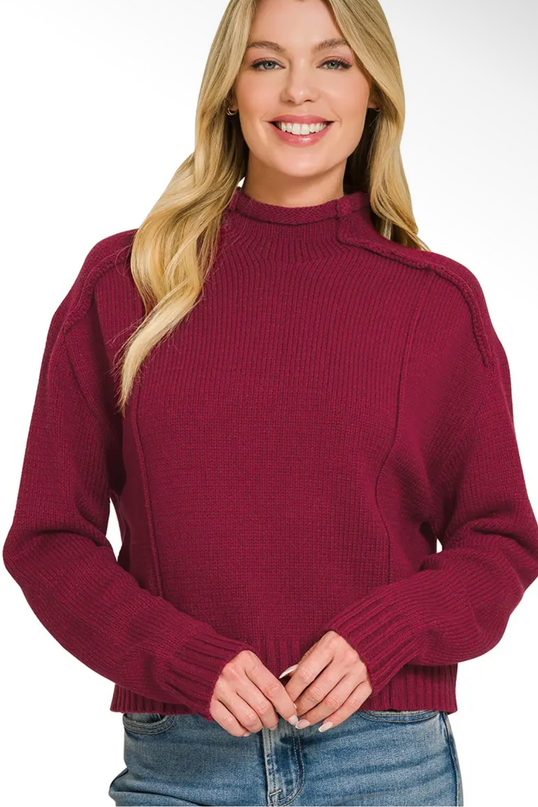 A woman wearing a ribbed mock neck sweater in burgundy, paired with blue jeans, shown against a plain white background.