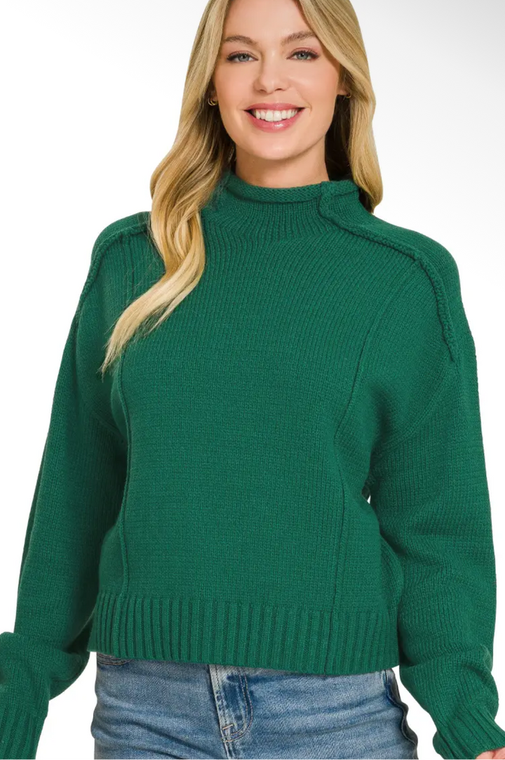 A woman wearing a ribbed mock neck sweater in green, paired with blue jeans, shown against a plain white background.