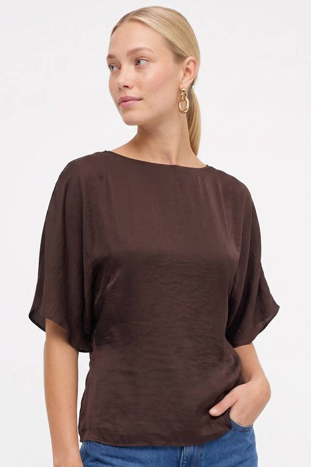 Woman wearing a chocolate brown silky dolman sleeve blouse with a boat neckline and blue jeans.